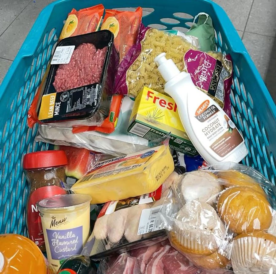 A shopping basket filled with various groceries, including ground beef, pasta, carrots, bread rolls, soap, custard, cheese, watermelon slices, and deli meats.