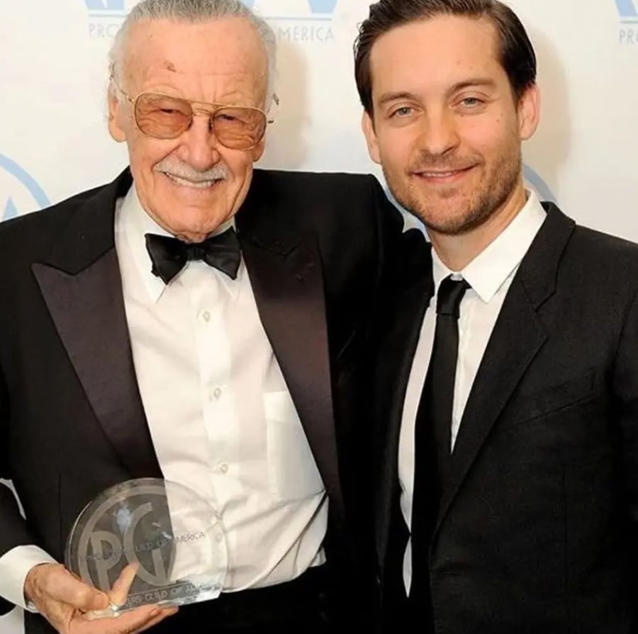 Two men wearing black suits and ties stand side by side, smiling at the camera. The older man on the left holds a glass award and wears sunglasses; the younger man on the right has his arm around his shoulder.