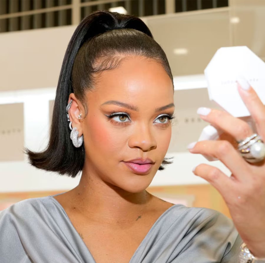 A woman with a sleek ponytail and elegant earrings holds a compact mirror, gazing at her reflection. She wears a gray top and has flawless makeup in a well-lit, modern setting.