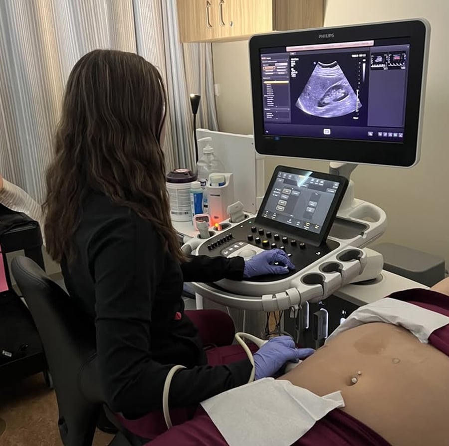 A medical professional performs an ultrasound on a person's abdomen. The patient lies on an exam table with their stomach exposed, while the technician uses a handheld device and monitors the ultrasound images on a nearby screen.