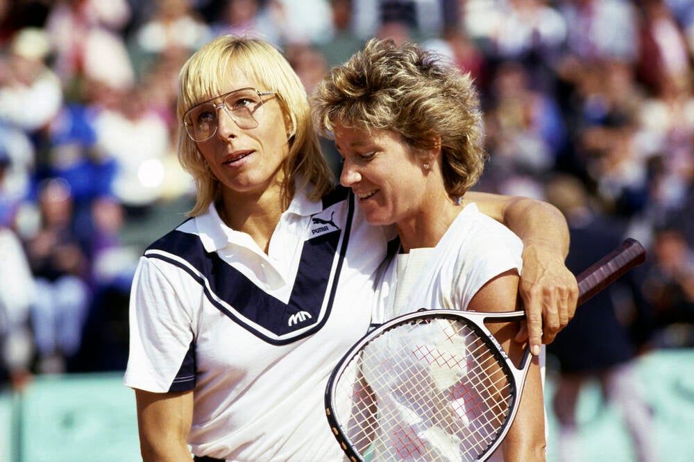 Two female tennis players stand side by side on a tennis court; one has her arm around the other. Both are smiling and holding tennis rackets, with a blurred crowd visible in the background.