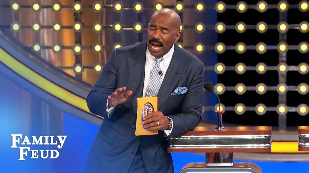 A man in a suit, holding a card, stands at a podium on the set of Family Feud, speaking animatedly. The Family Feud logo is visible in the bottom left corner, and round lights decorate the background.