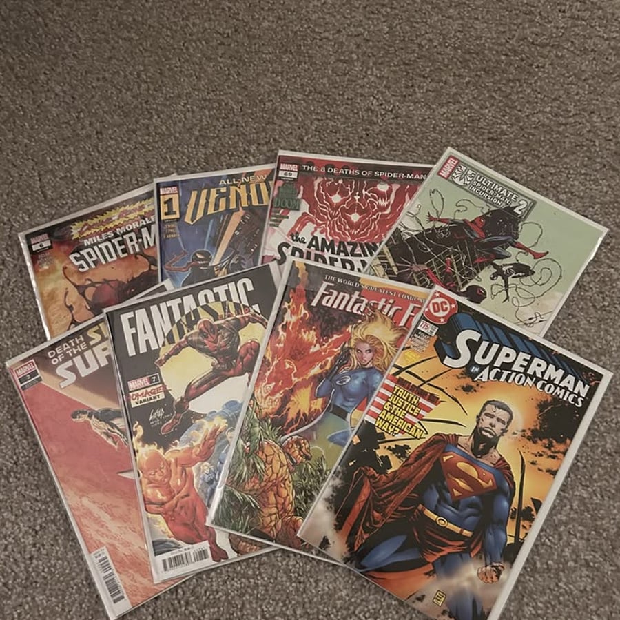 Eight comic books are laid out on a carpeted floor, featuring titles like Spider-Man, Venom, Fantastic Four, and Superman Action Comics with colorful, dynamic cover art.