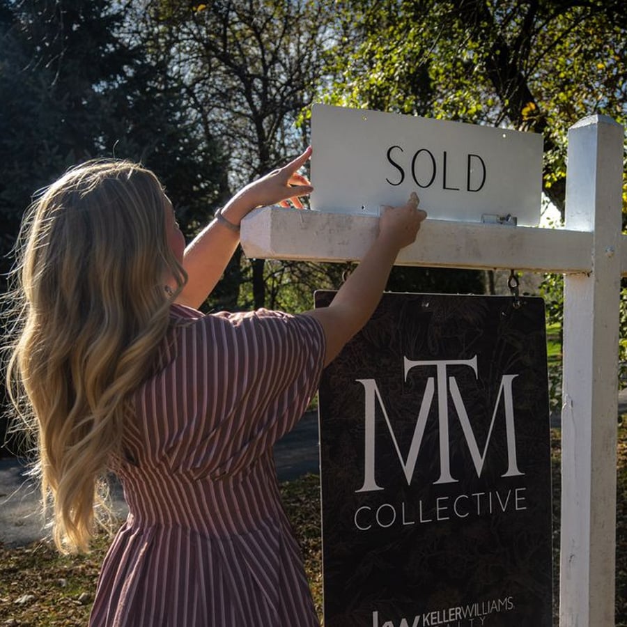 A woman with long blonde hair places a "SOLD" sign on a white real estate post, beneath which hangs a sign for MTM Collective. Trees and sunlight are visible in the background.