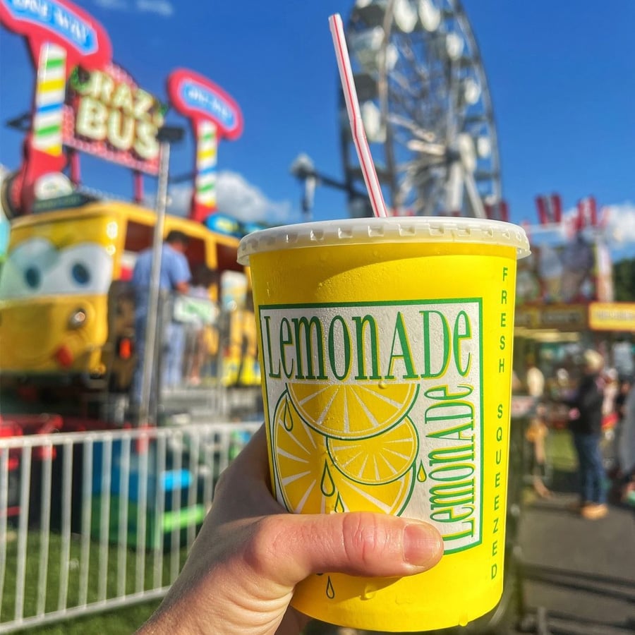 A hand holds a yellow cup labeled “Lemonade” with a red-and-white striped straw. In the background, there is a carnival scene with a ferris wheel and colorful rides, including one shaped like a bus.