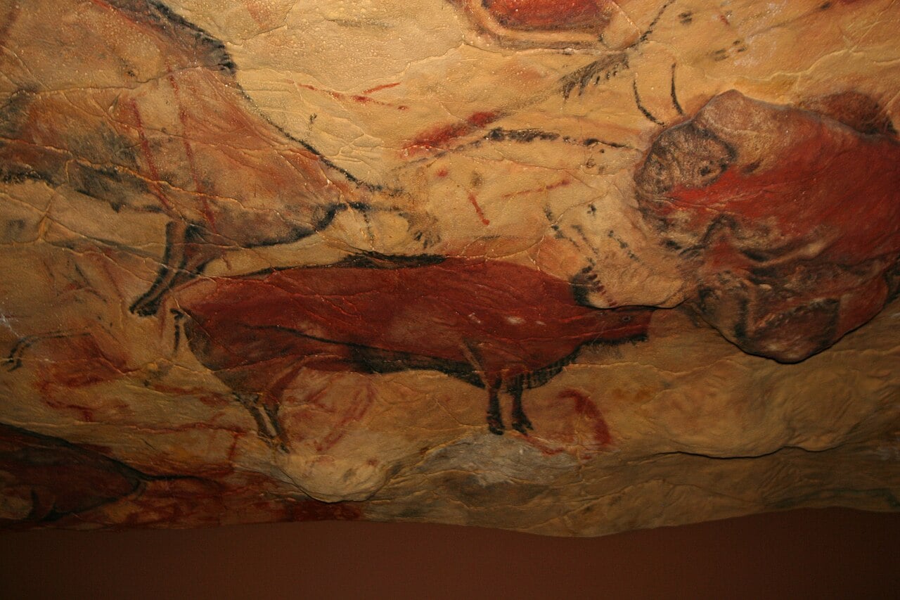 Cave painting depicting bison in shades of red and brown with dark outlines, on a textured rock surface with cracks and natural color variations.