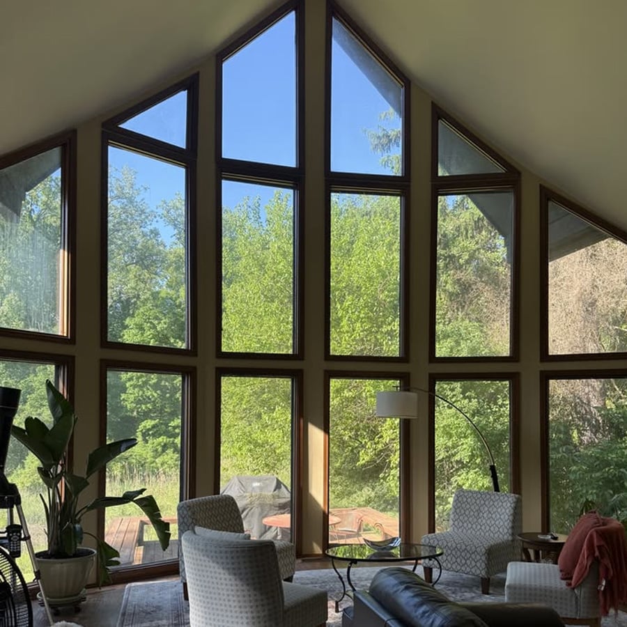 A living room with tall, triangular windows offers a view of lush green trees outside. Two patterned chairs, a round table, a floor lamp, and a potted plant are arranged near the windows, letting in abundant natural light.