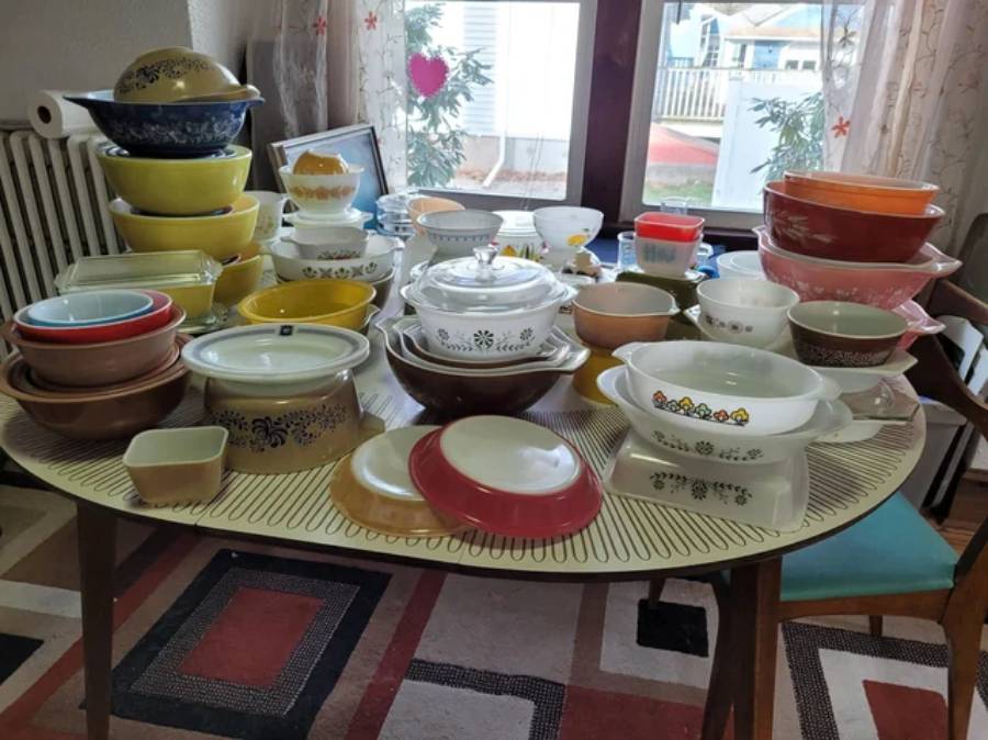 A large collection of colorful vintage Pyrex dishes, bowls, and containers arranged on a dining table, with a patterned rug underneath and sunlight filtering through a window in the background.