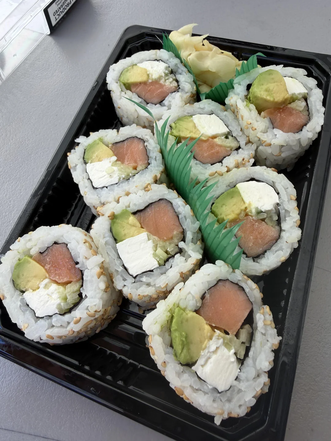 A black plastic tray with eight pieces of sushi rolls filled with salmon, avocado, and cream cheese, garnished with pickled ginger and plastic grass dividers.