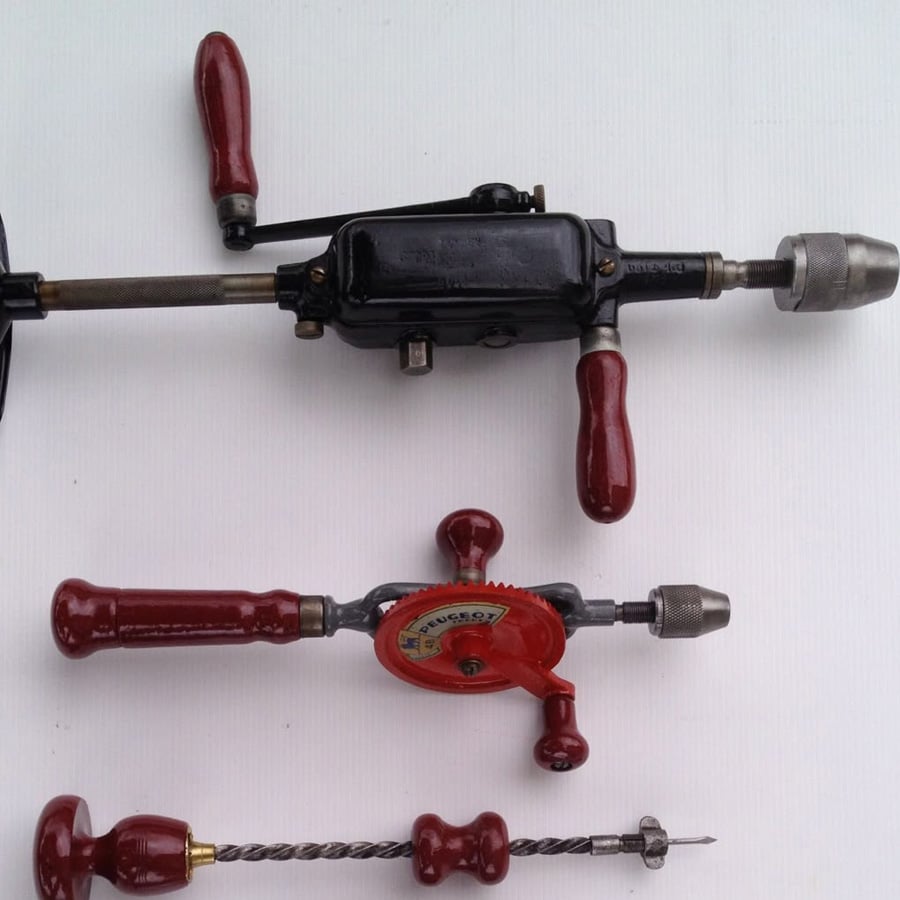 Three vintage hand drills with red handles are arranged on a white background, including a black hand-crank drill at the top, a red geared hand drill in the middle, and a wooden-handled brace drill at the bottom.