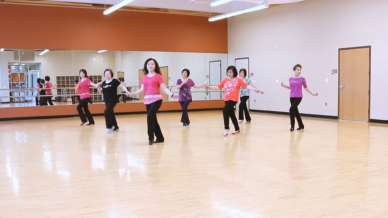 A group of women in colorful shirts and black pants practice a dance routine in a spacious, mirrored studio with wooden floors and bright lighting.