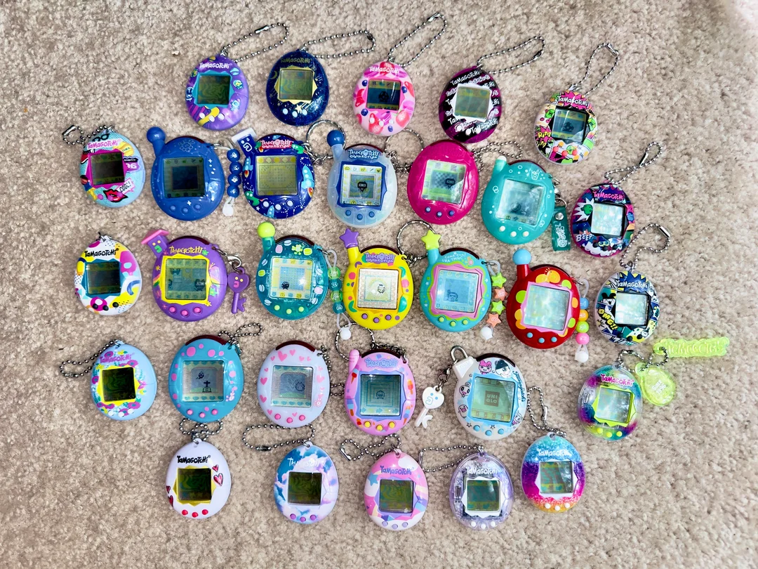 A collection of colorful Tamagotchi virtual pets arranged in rows on a beige carpet, featuring various shapes, patterns, and character designs with attached keychains.