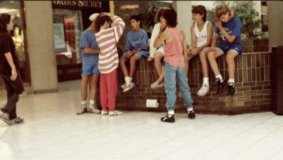 A group of teenagers in casual 80s or 90s clothing gather and chat inside a shopping mall, some sitting on a brick ledge, with stores and shoppers in the background.