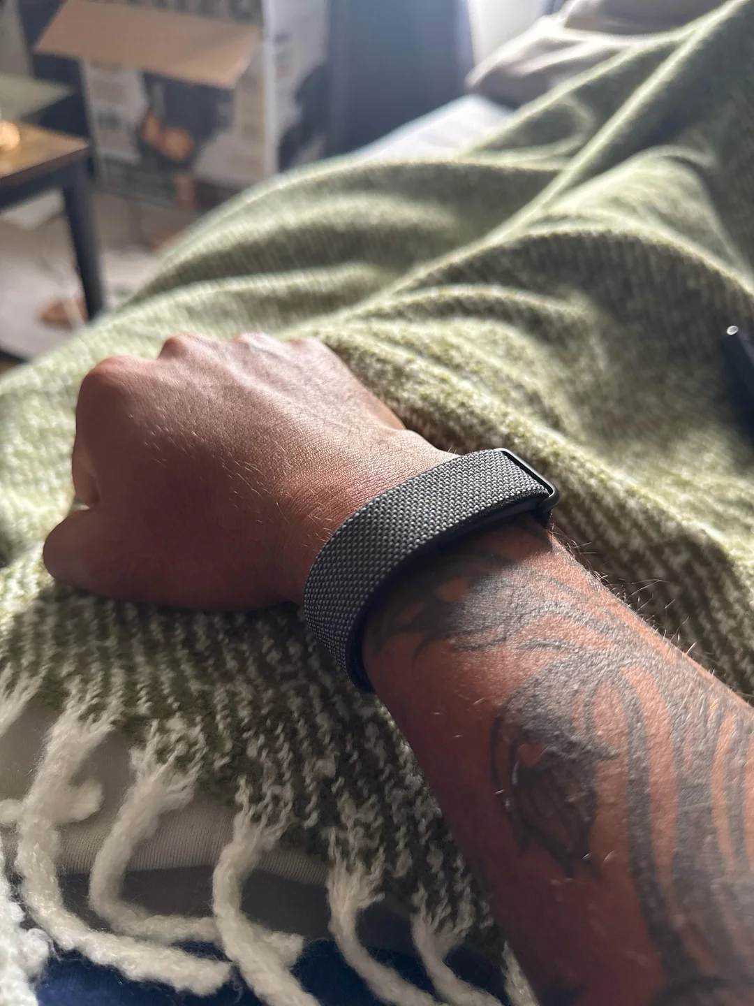 A tattooed arm with a smartwatch rests on a green and white fringed blanket. The background shows a slightly blurred, open cardboard box and some household items.
