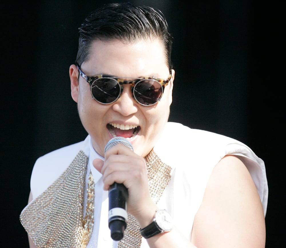 A man wearing sunglasses and a sleeveless white and gold outfit is singing into a microphone. He has slicked-back dark hair and is performing on stage.