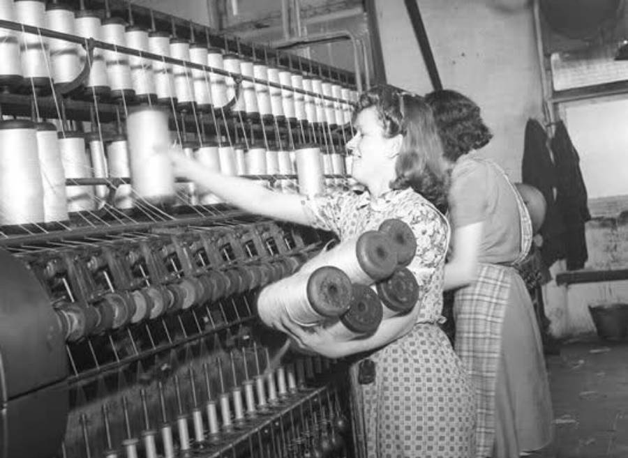 Two women work in a textile factory, handling large spools of thread on an industrial machine. One woman reaches to place or retrieve a spool, while the other operates at a different section of the machine.