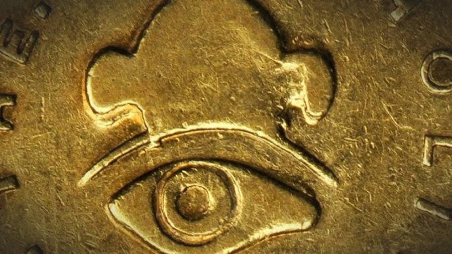 A close-up of a gold coin shows an embossed design featuring an eye with a stylized hat or abstract shape above it, surrounded by engraved markings and textured surface details.