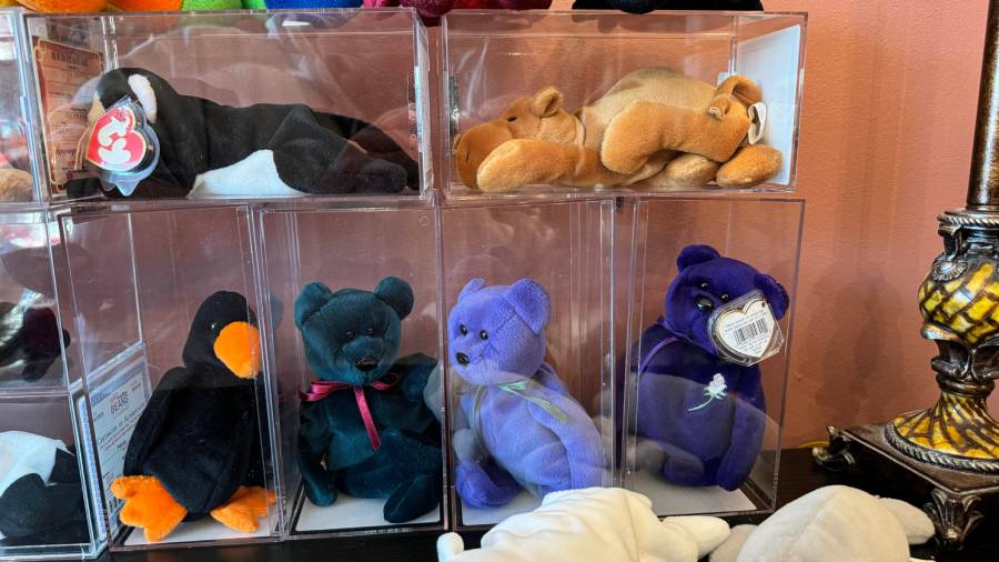 A collection of Beanie Babies, including a penguin, a bear, and a camel, displayed in clear plastic boxes on a shelf next to a lamp with a yellow patterned base.