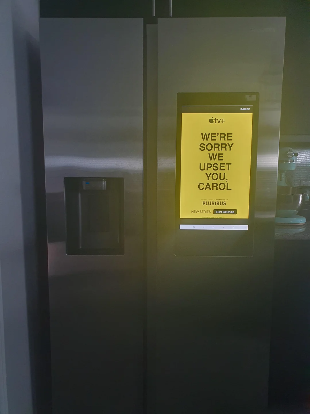 A refrigerator with a built-in screen displaying a bright yellow Apple TV+ ad reading, "WE'RE SORRY WE UPSET YOU, CAROL" in bold black text. The fridge is in a modern kitchen setting.