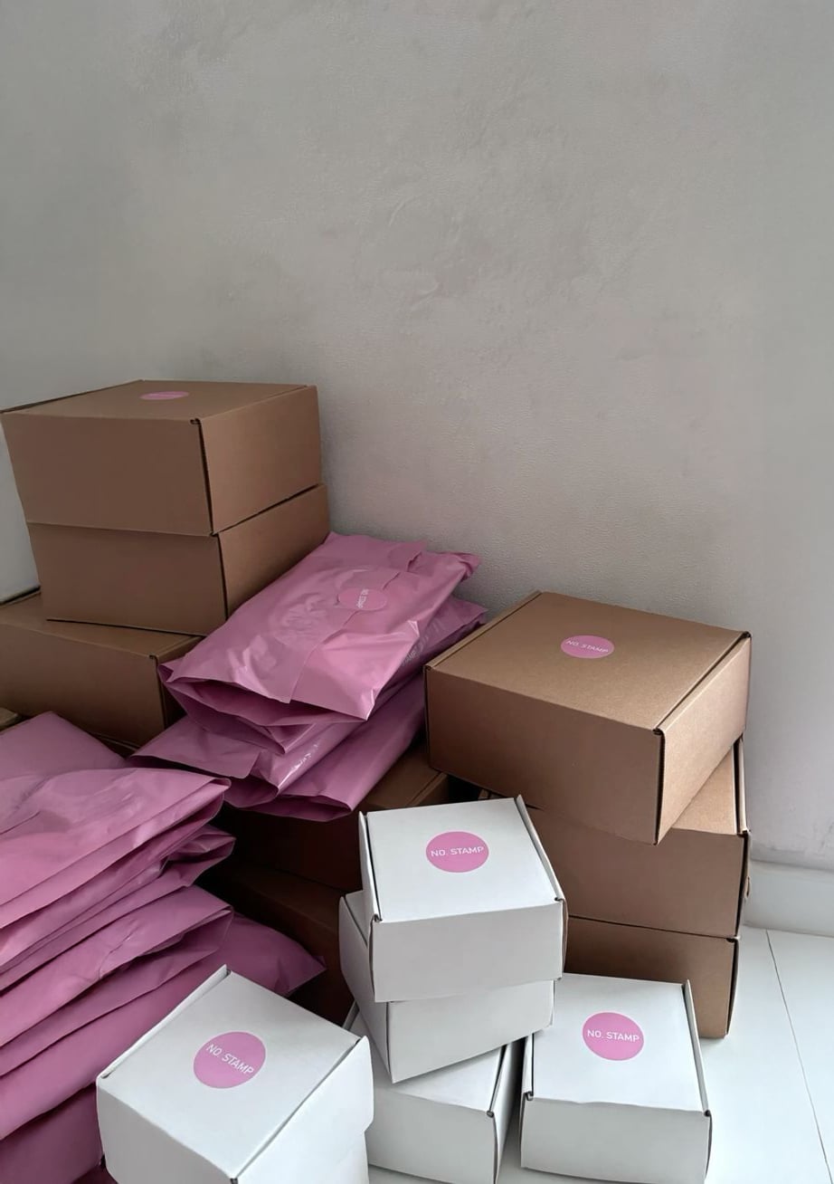 A stack of brown cardboard boxes, white boxes, and pink shipping bags, each with a pink sticker, are arranged against a plain white wall on a white tiled floor.
