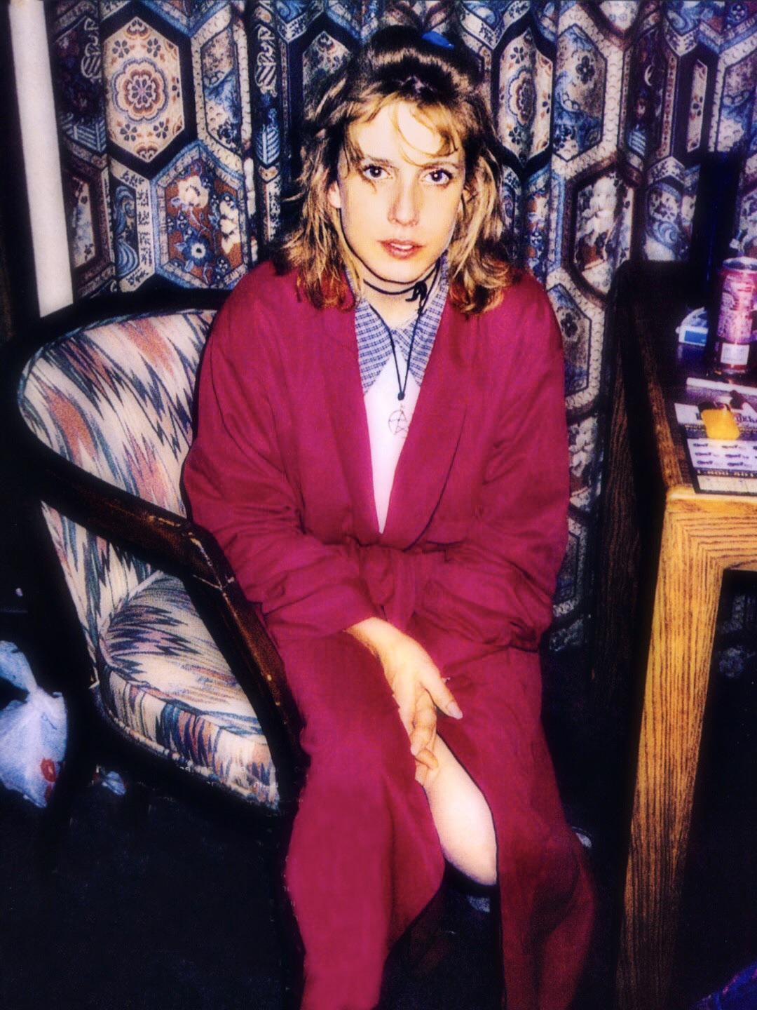 A woman with blonde hair sits on a patterned chair, wearing a red robe and a necklace. She looks at the camera. Behind her are patterned curtains, a table with various items, and a trash bag on the floor.