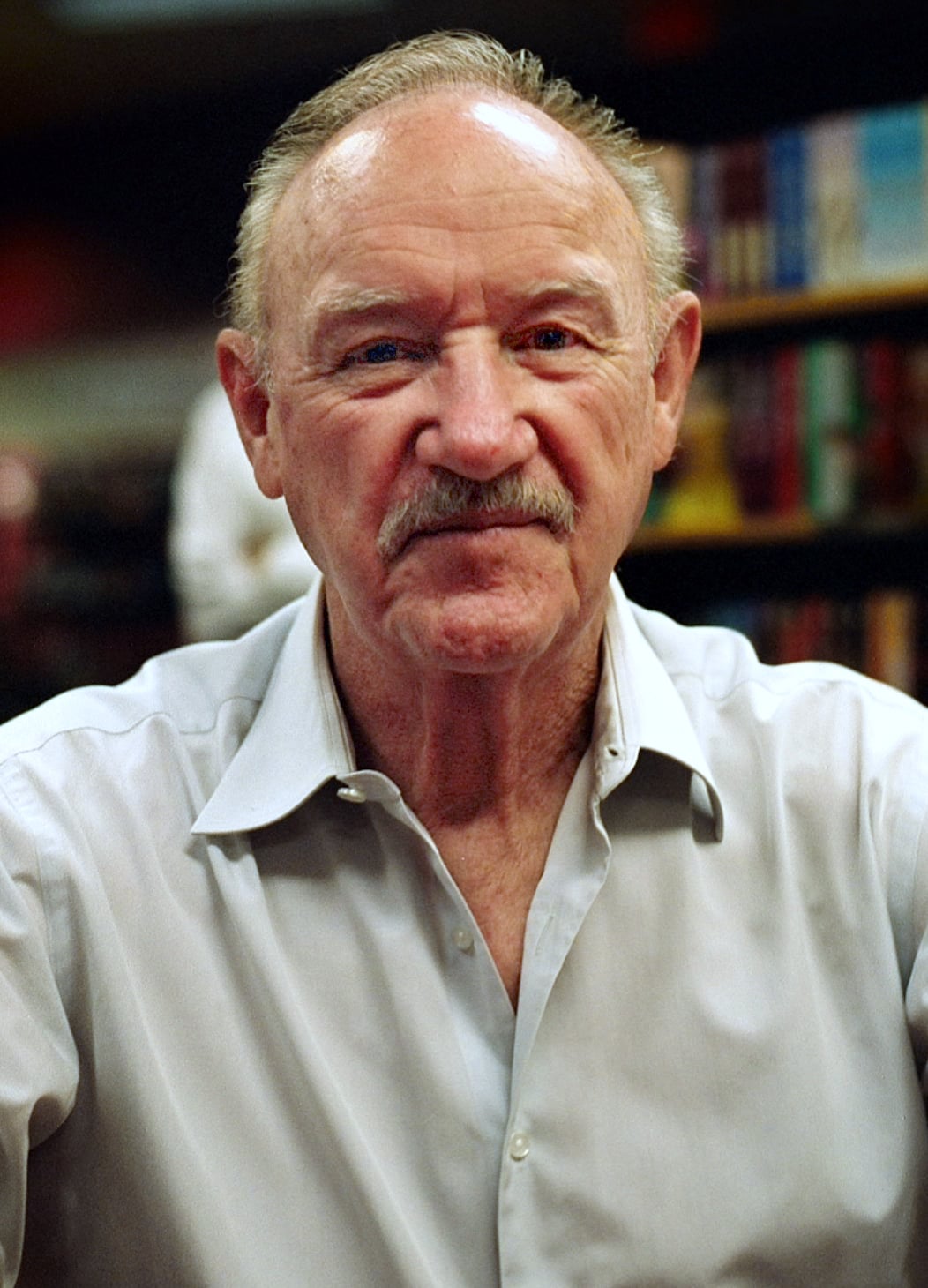 An older man with a mustache and short hair, wearing a light gray button-up shirt, sits indoors with shelves of books blurred in the background.