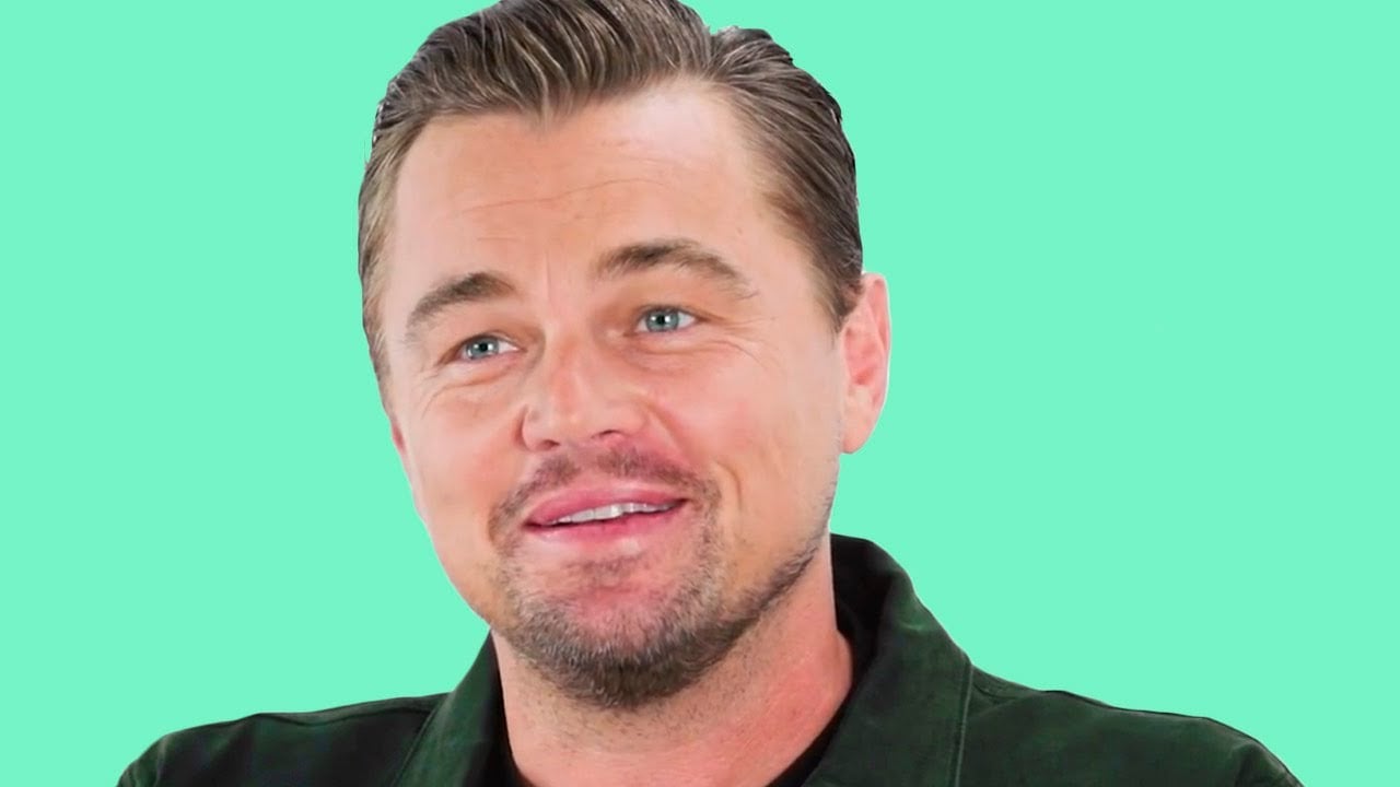A smiling man with short, slicked-back hair, light stubble, and blue eyes, wearing a dark green jacket, in front of a solid light turquoise background.