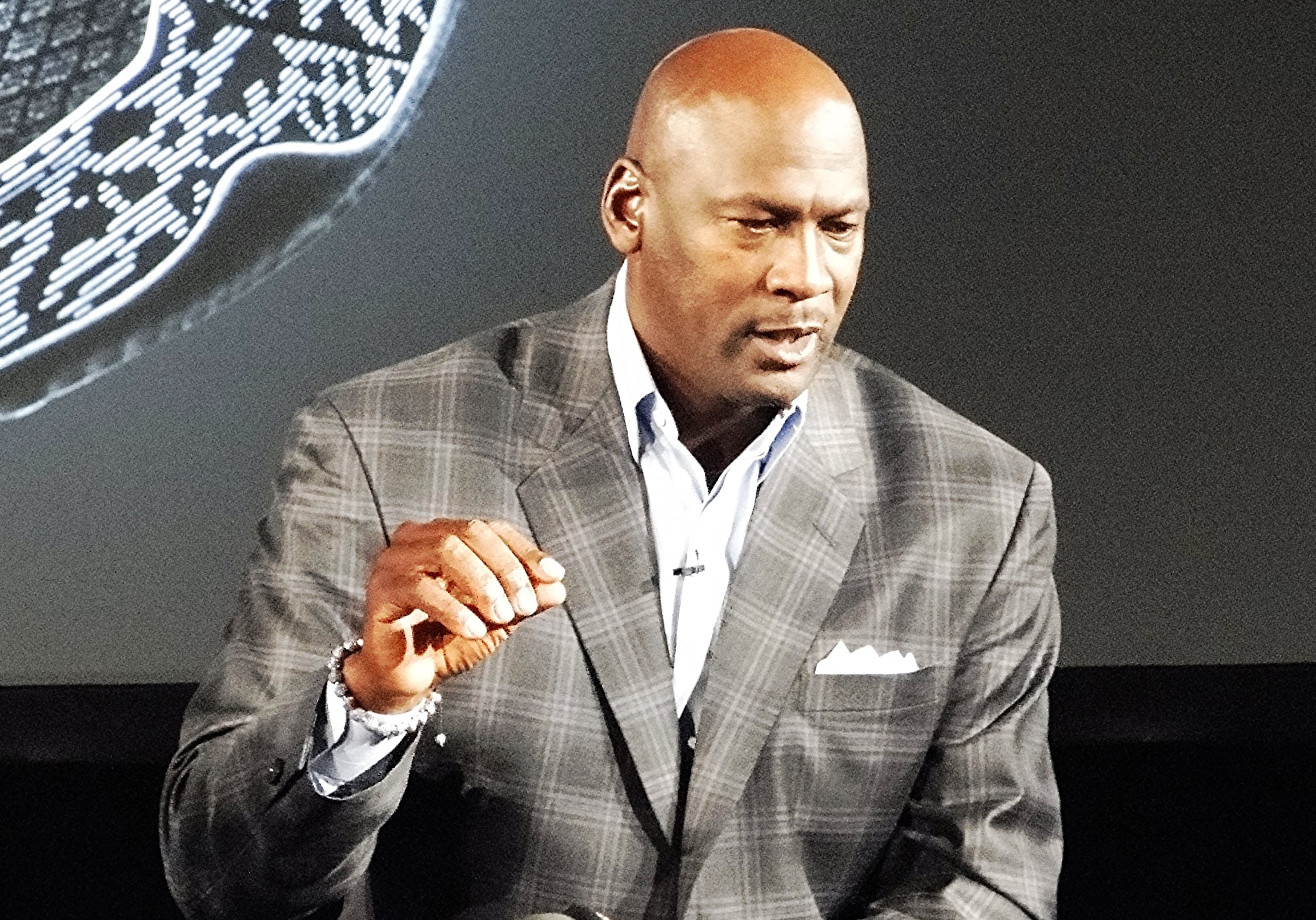 A man in a checked suit jacket and light blue shirt speaks while gesturing with his right hand. He is sitting in front of a dark background with a patterned object partially visible.