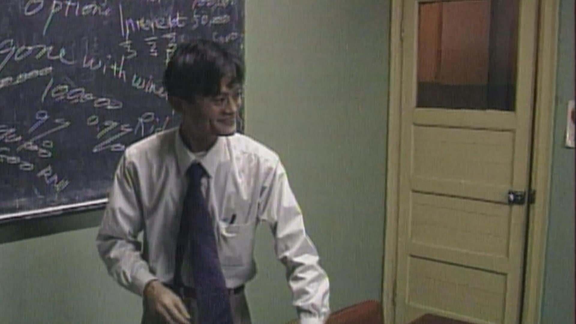 A man in a white shirt and tie stands in a classroom near a chalkboard covered with handwritten notes and a closed yellow door.