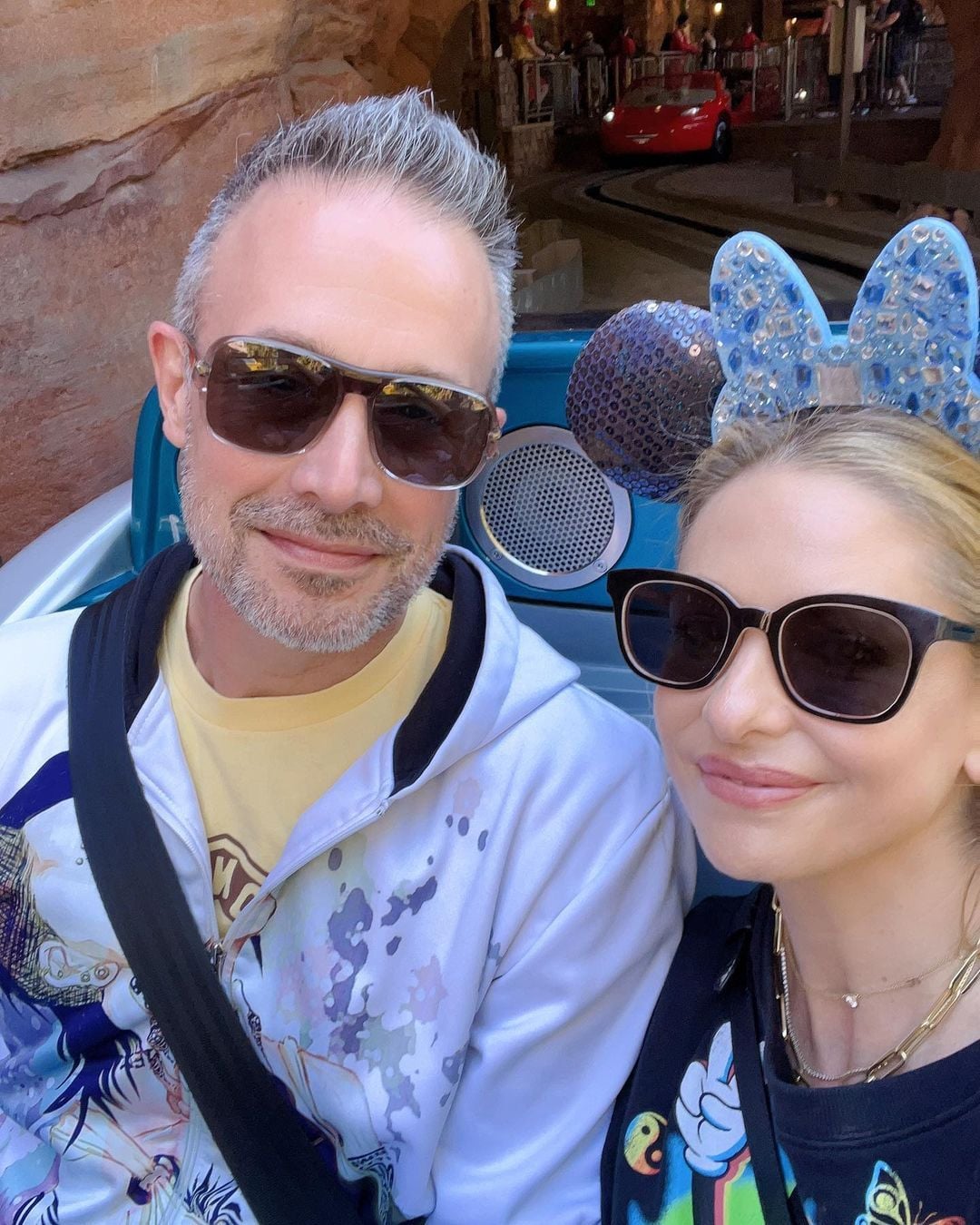 A man and a woman wearing sunglasses smile for a selfie on an amusement park ride. The woman has sparkly blue Minnie Mouse ears, and a red car is visible in the background.