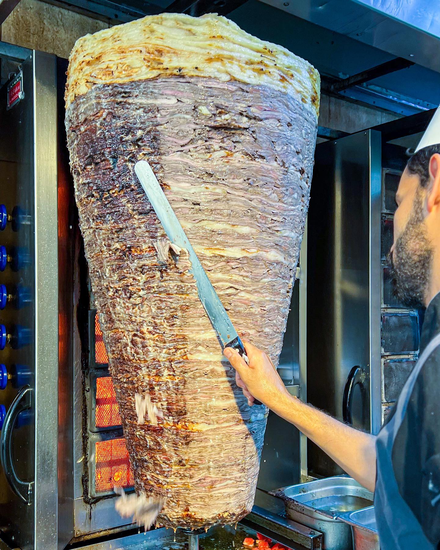 A person slices meat from a large vertical rotisserie shawarma spit in a restaurant kitchen, using a long, thin knife to carve thin pieces as the meat cooks beside an open flame.