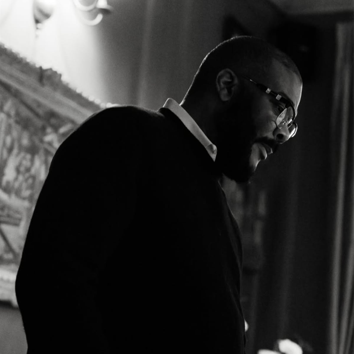 A man in glasses and a sweater stands indoors, looking downward in a dimly lit room with framed art on the wall behind him. The photo is in black and white.