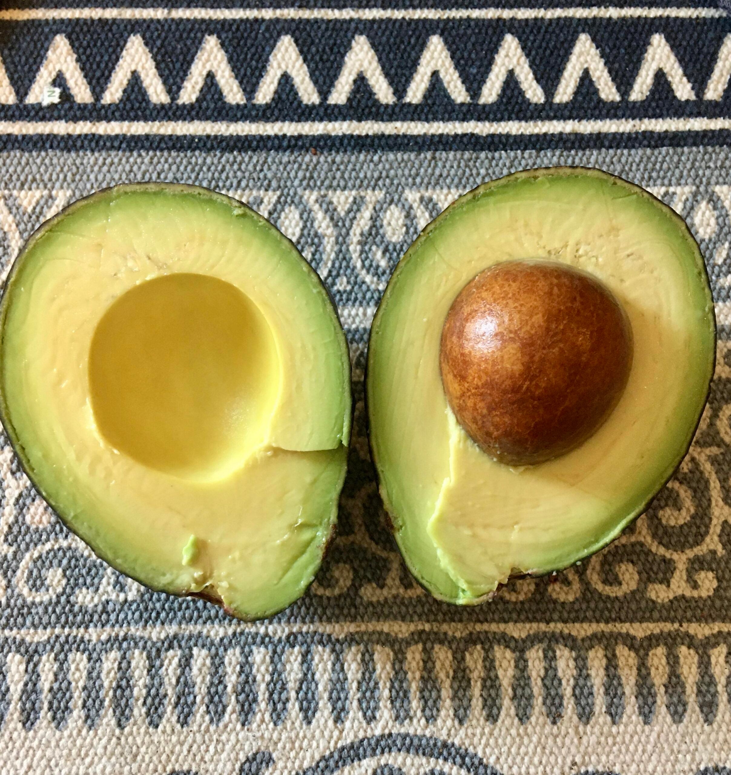 A ripe avocado cut in half, with one half showing the seed and the other half empty, placed on a blue and white patterned fabric.