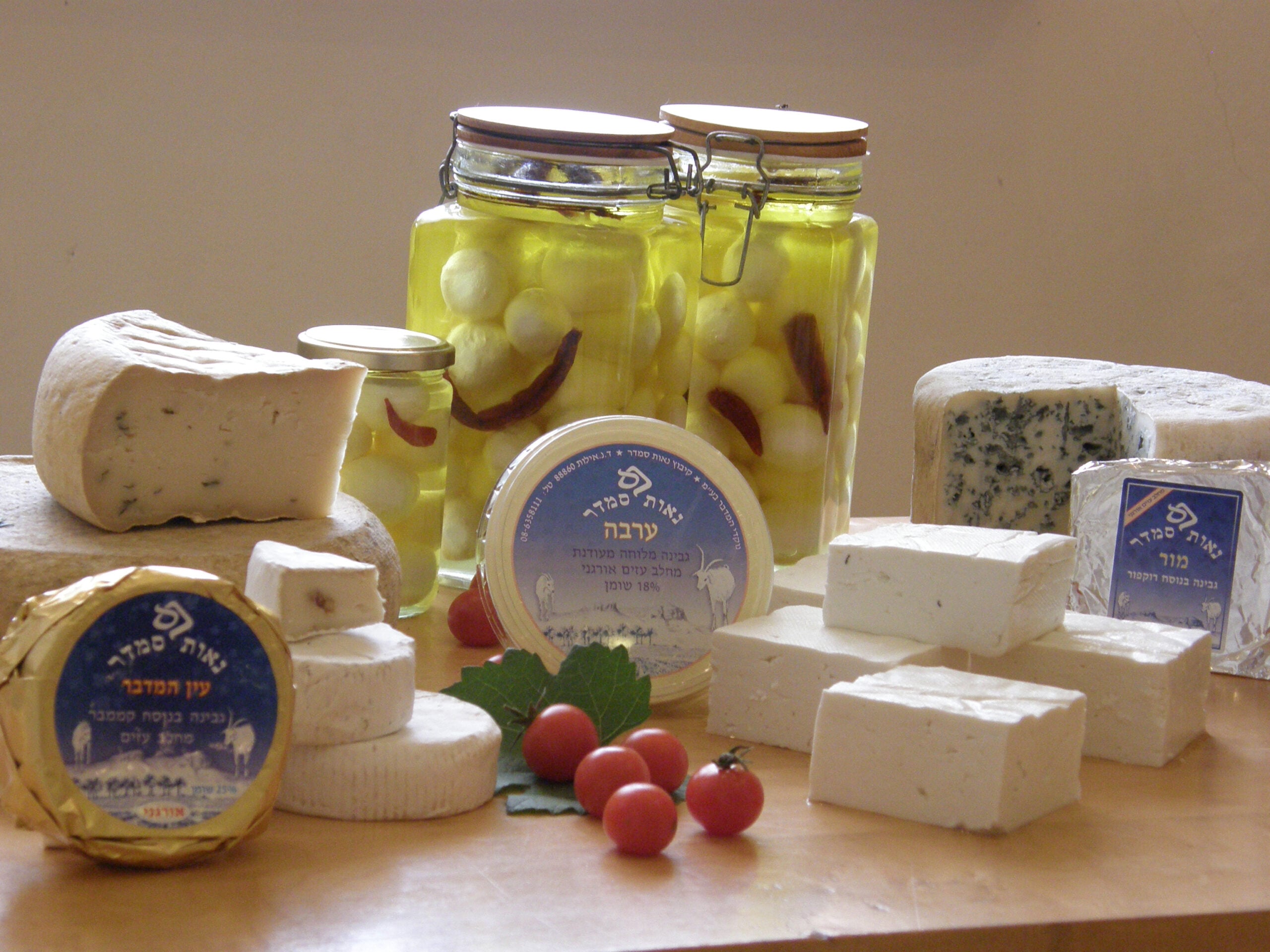 An assortment of cheeses, including round, block, and wedge shapes, some wrapped and labeled, arranged with jars of cheese in oil, cherry tomatoes, and a green leaf on a wooden surface.