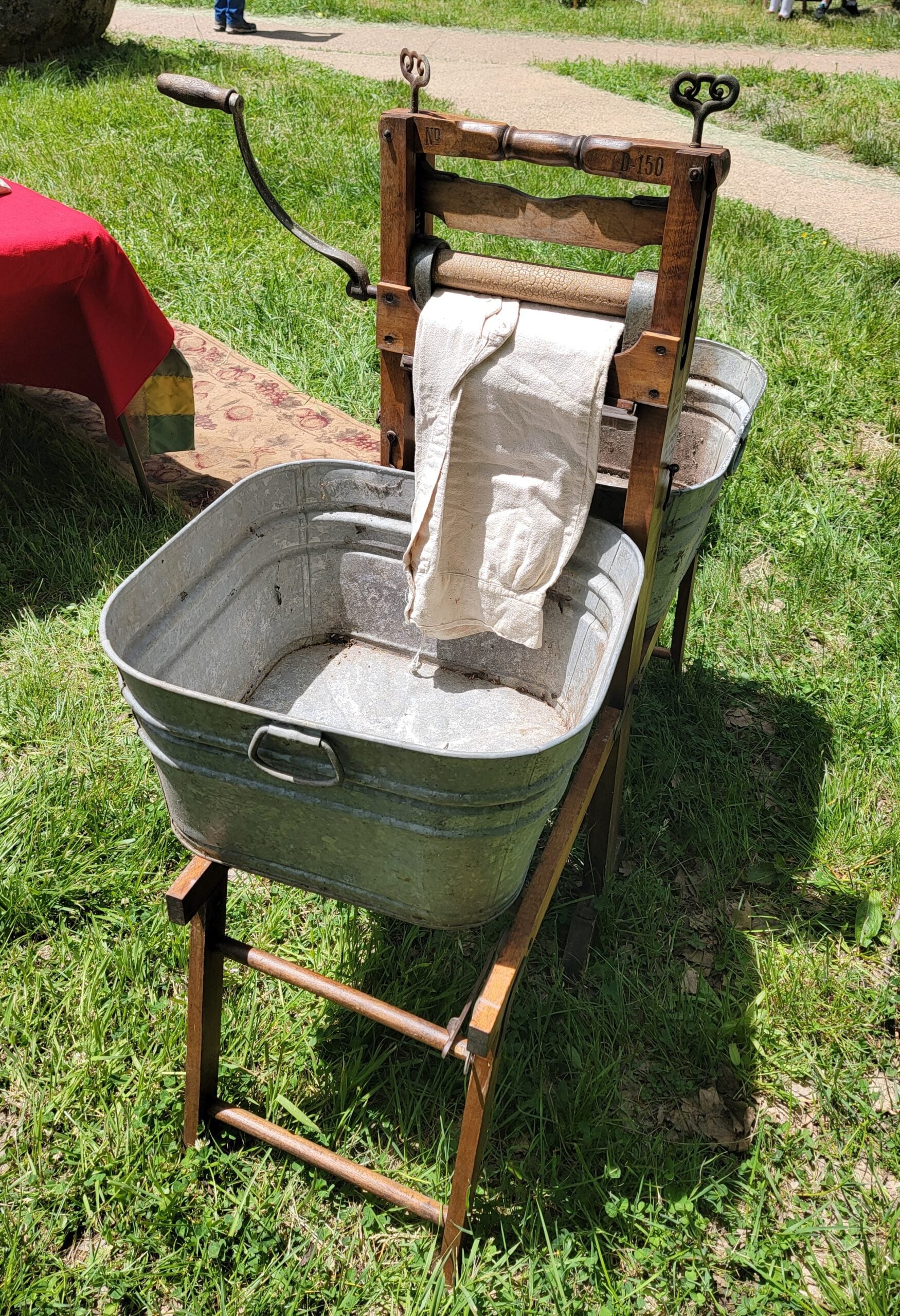A vintage manual washing machine with two metal tubs and a hand-cranked wringer holds a wet cloth draped over the rollers, set outdoors on grass under sunlight.