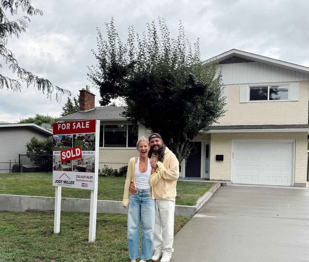 A smiling couple stands in front of a house with a "For Sale" sign that reads "Sold." They appear happy, embracing each other on the driveway. The house is two stories with a white garage and a well-kept yard.