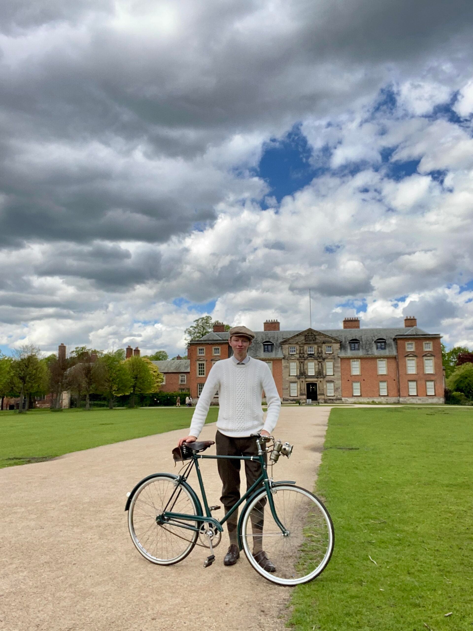 A man in vintage-style clothing stands holding a bicycle on a path, with a large historic mansion and cloudy sky in the background, surrounded by green grass and trees.