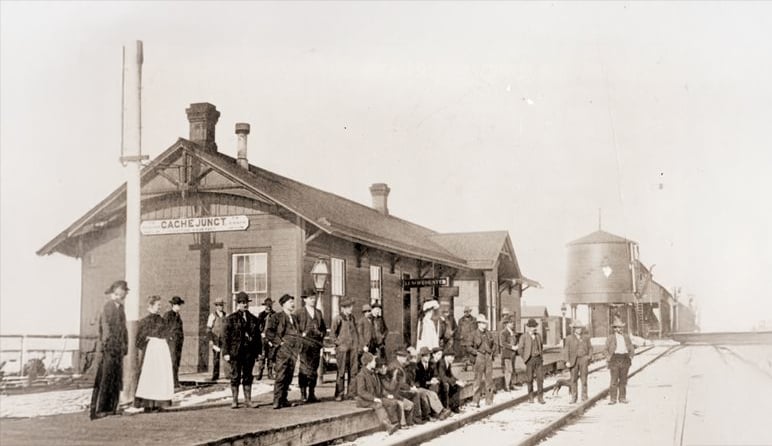 A group of people, including men, women, and children, stands and sits on the platform outside a small train station labeled “Cache Junction.” A water tower and train tracks are visible on the right. The photo is in black and white.