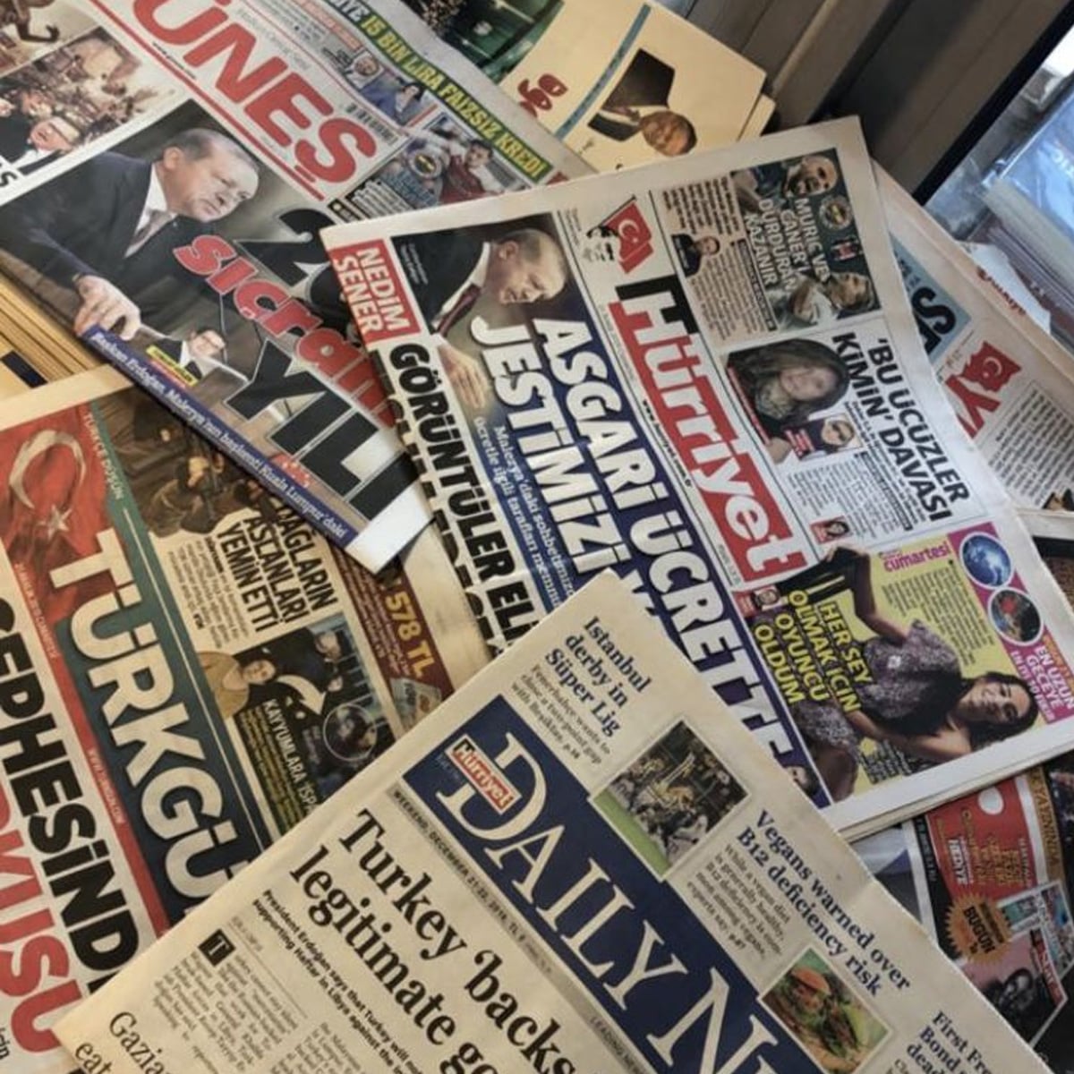 Several Turkish and English-language newspapers spread out on a table, showing headlines, photos of people, and colorful text in Turkish and English. The newspapers are overlapping and include Hürriyet and Daily News.