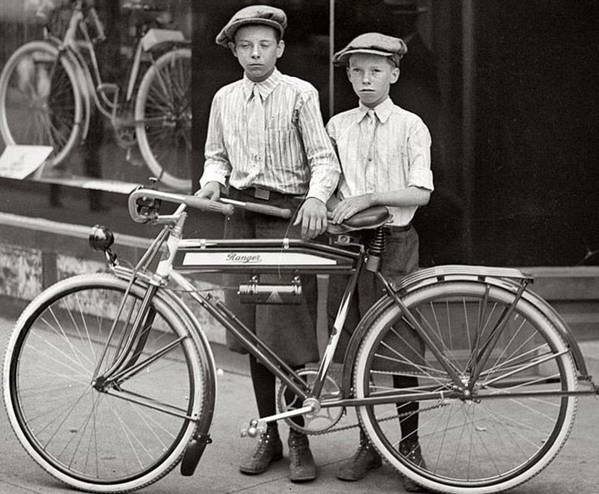 Two boys wearing flat caps and striped shirts stand beside a vintage bicycle on a city sidewalk, with a store window displaying more bicycles behind them.