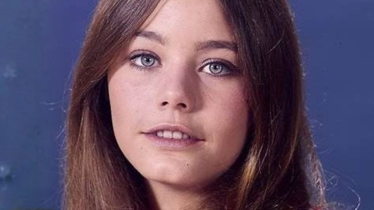 A young woman with long brown hair and blue eyes looks at the camera, smiling softly. She is in front of a plain blue background.
