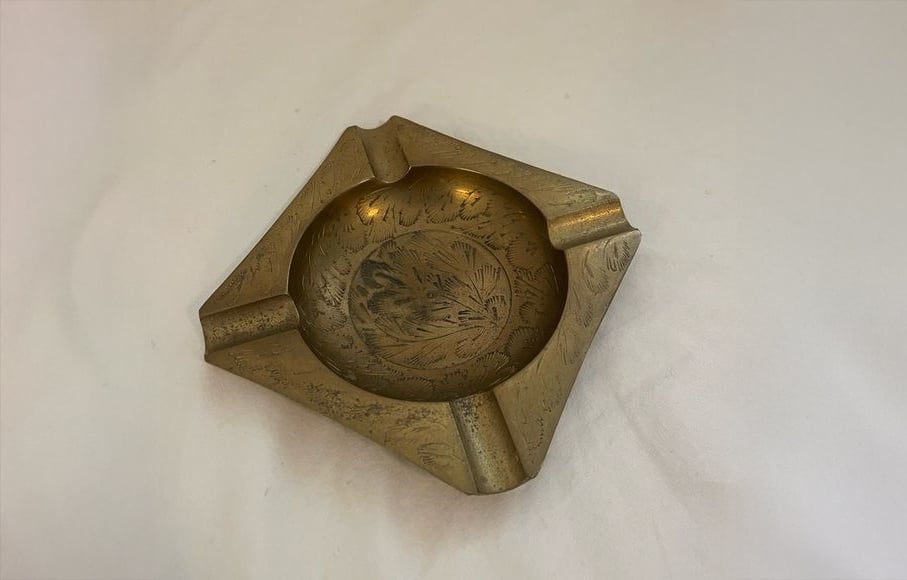 A brass ashtray with engraved floral patterns, featuring a round bowl and three angular cigarette rests, placed on a white surface.