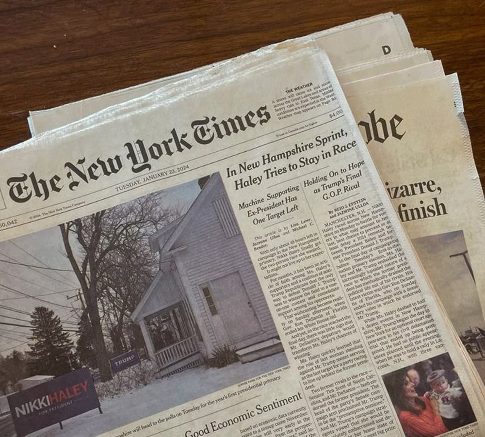 A close-up of two overlapping newspapers on a table, with The New York Times on top. The visible headline reads, "In New Hampshire Sprint, Haley Tries to Stay in Race." An image of a house with a Trump sign is shown.