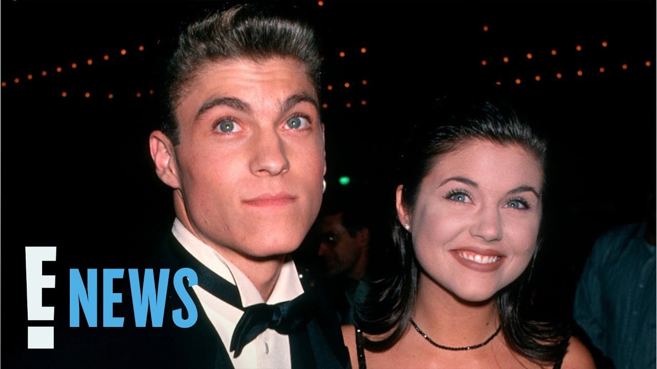 A young man in a tuxedo and a young woman in a black dress smile at an event, with string lights in the background. The "E! News" logo appears in the corner.