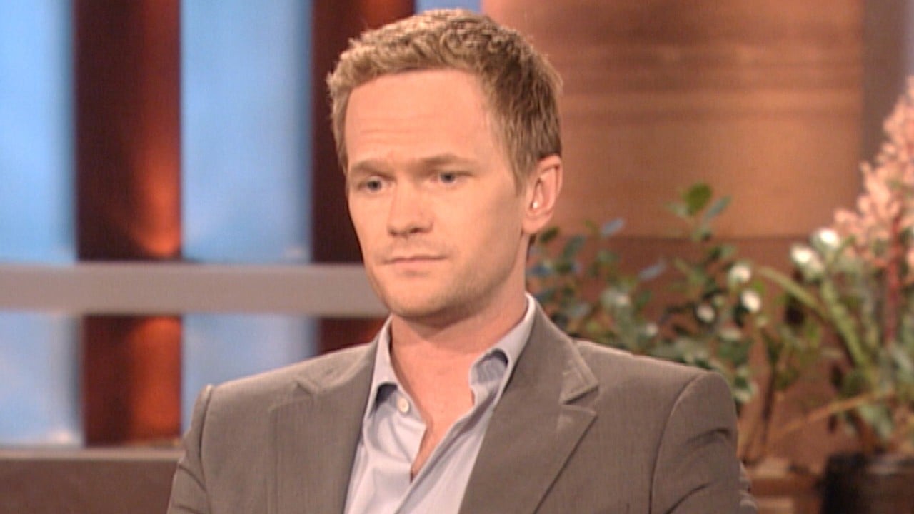 A man in a gray suit with short light brown hair sits indoors, looking serious and thoughtful. There are blurred plants and warm-toned decor in the background.