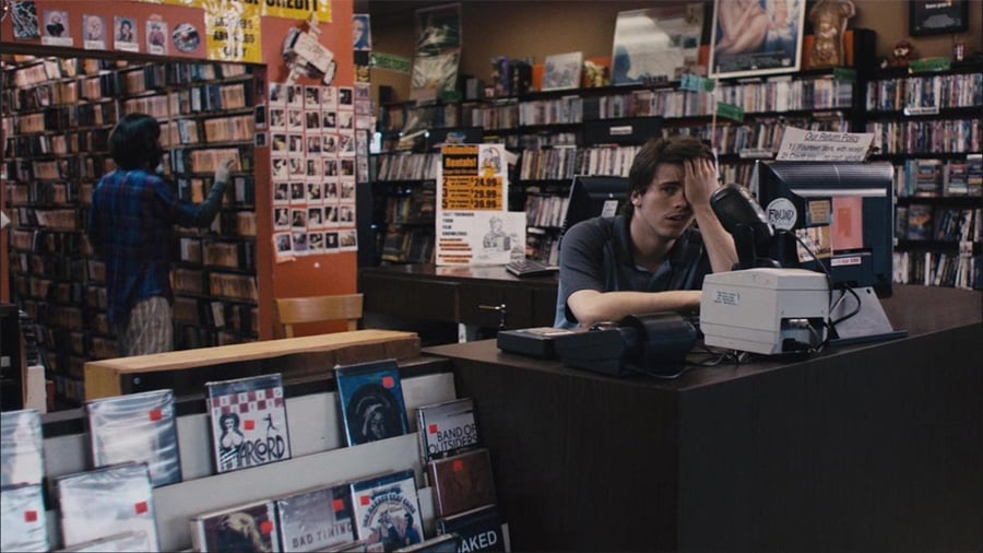 A young man sits behind the counter of a cluttered video rental store, looking bored with his hand on his forehead. Rows of DVDs and CDs fill the shelves, and another person browses in the background.