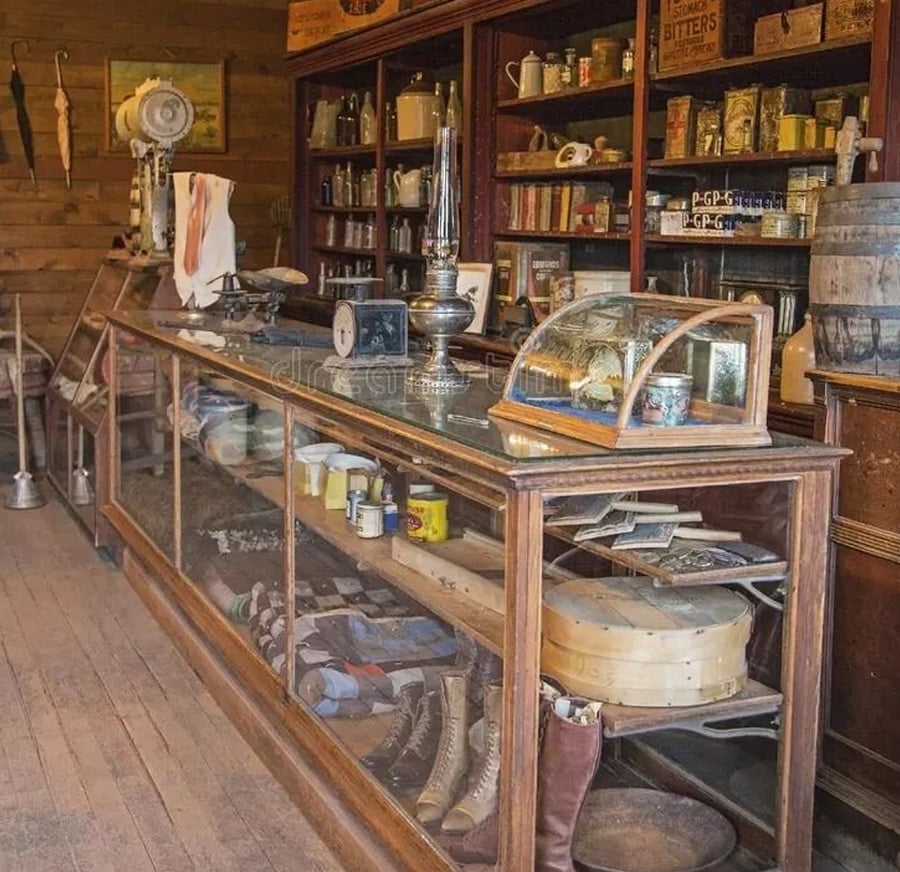 An old-fashioned general store with wooden shelves stocked with jars, bottles, and tins. Glass display cases hold boots, hats, and tools. A vintage scale and oil lamp sit on the counter, giving a rustic, historical feel.