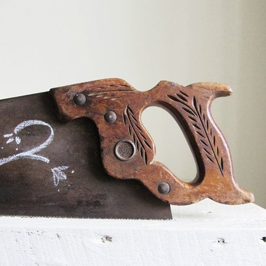 A vintage hand saw with a wooden handle featuring engraved leaf designs rests on a white surface. The blade has chalk markings, including the number 2 and an arrow pointing left.