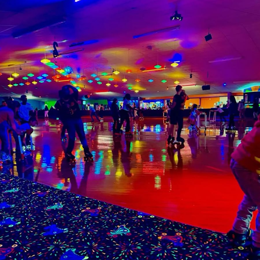 People are roller skating in a rink lit with colorful neon and black lights, creating vibrant reflections on the floor. The scene is lively, with groups of people skating and socializing under bright, colorful ceiling lights.