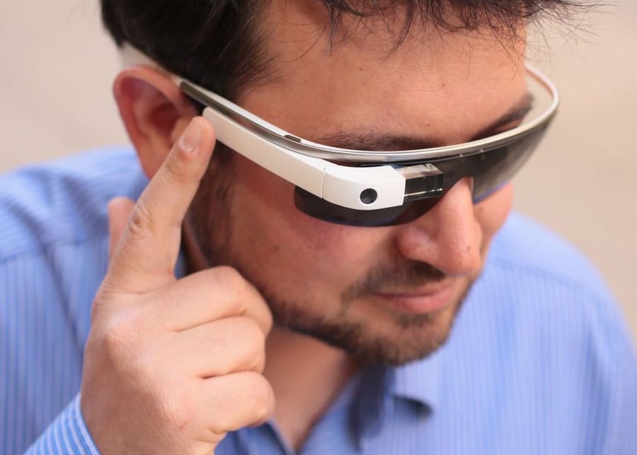 A man wearing a blue shirt uses a pair of smart glasses, touching the side of the device with his finger to interact with it.